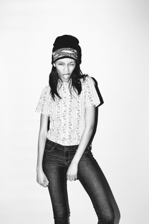 Young woman wearing a beanie and patterned t-shirt posing playfully against a white wall