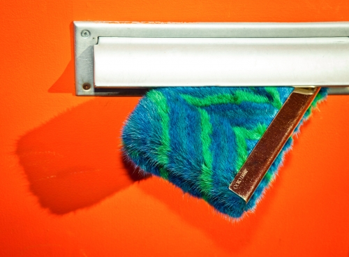 A fuzzy blue and green striped object is coming out of a silver mail slot on an orange door, with a visible shadow.