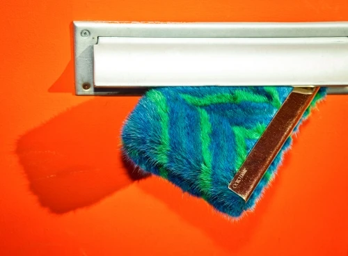 A fuzzy blue and green striped object is coming out of a silver mail slot on an orange door, with a visible shadow.