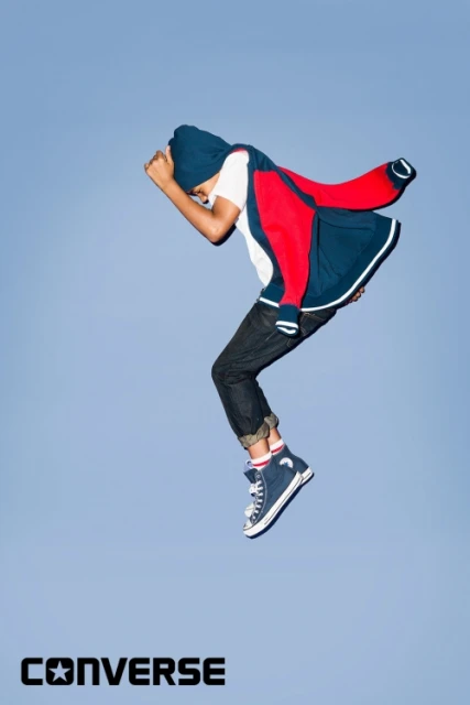 Young child in mid-air wearing jacket, jeans, and blue hi-top Converse sneakers against a clear blue sky.