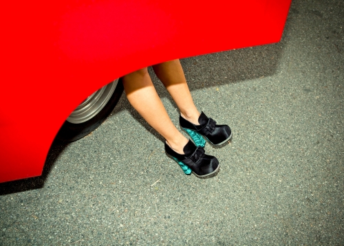 Woman's legs extend from under a red car, feet adorned with satin shoes with a teal wedge heel, set on asphalt.