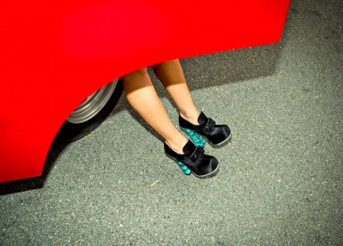 Woman's legs extend from under a red car, feet adorned with satin shoes with a teal wedge heel, set on asphalt.