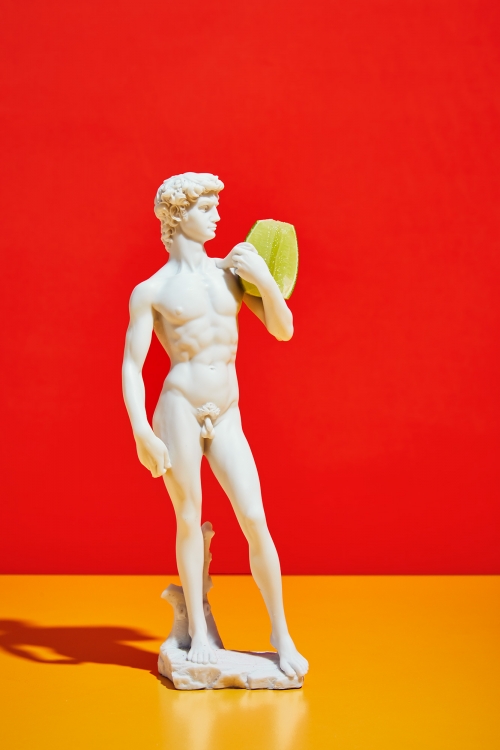 Photograph of a David statue holding a loofah against a red and yellow background. The statue is made of white stone.