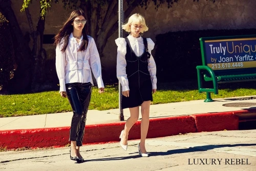 Two women walk on a sunny street in fashionable outfits, showcasing modern style and urban flair with a bench and greenery in the background.