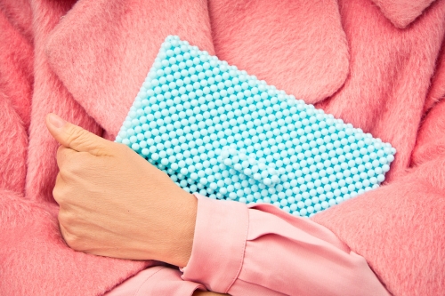 Hand holds a light blue beaded clutch against a backdrop of a pink faux fur coat, with a pink shirt sleeve visible.
