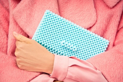Hand holds a light blue beaded clutch against a backdrop of a pink faux fur coat, with a pink shirt sleeve visible.