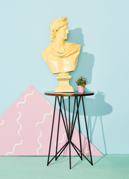Bust sculpture of male figure on round table with geometric shapes and mint green background in a studio-lit interior shot.