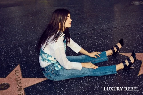 Brunette woman in jeans and heels poses on Hollywood Walk of Fame star, wearing a white shirt and floral vest in an outdoor setting.
