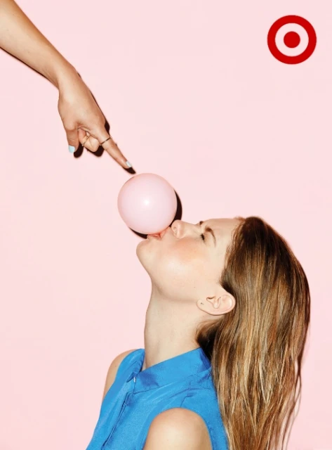A young woman with long hair holds a pink bubblegum bubble as another hand is about to pop the bubble in front of a pink background.