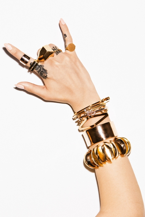 Close-up of a woman's hand adorned with gold rings and bracelets, making a 'rock on' gesture against a plain white backdrop.