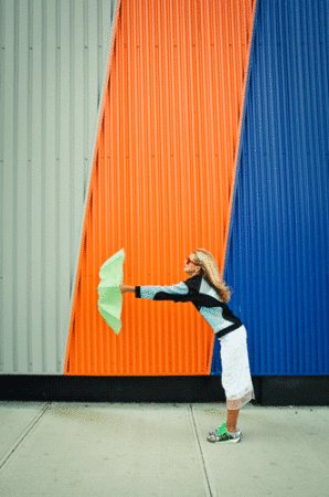 Woman leaning forward holding a green paper fan against a corrugated metal wall with diagonal stripes of grey, orange, and blue.