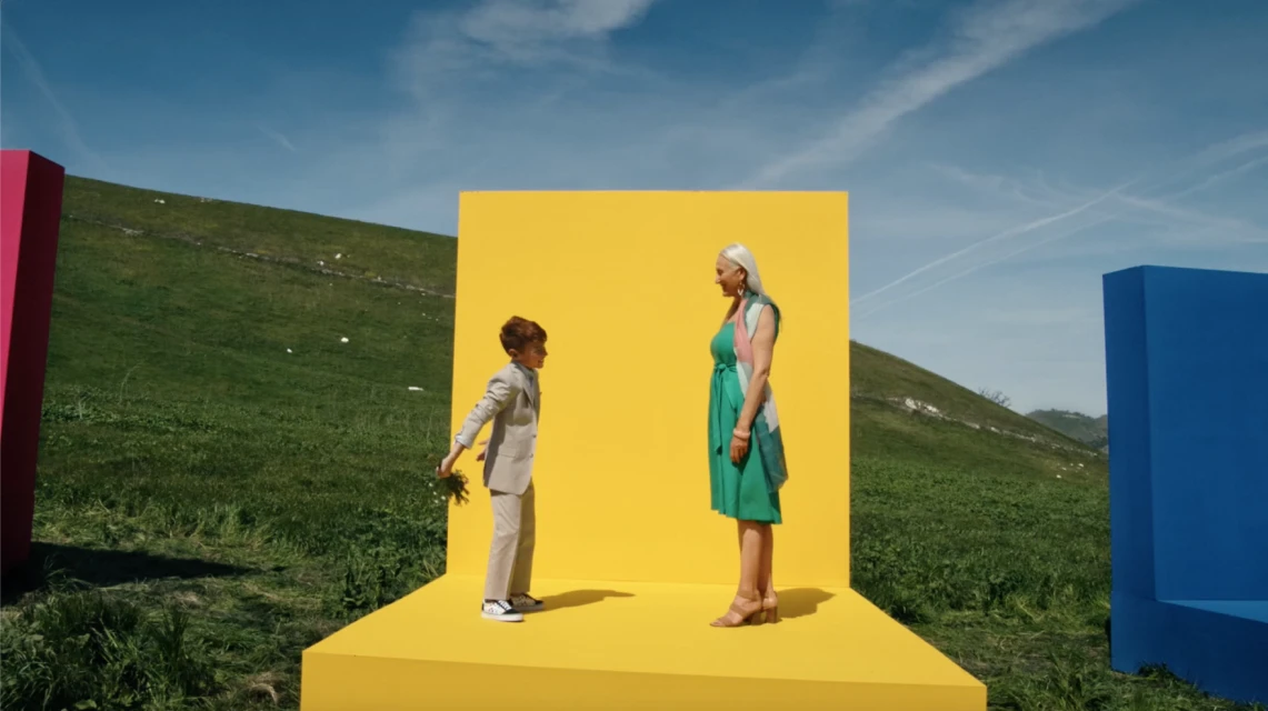 Boy in suit hands flowers to woman in green dress in outdoor setting with colorful geometric shapes under blue sky.