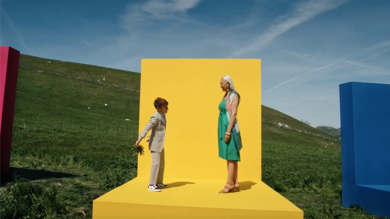 Boy in suit hands flowers to woman in green dress in outdoor setting with colorful geometric shapes under blue sky.