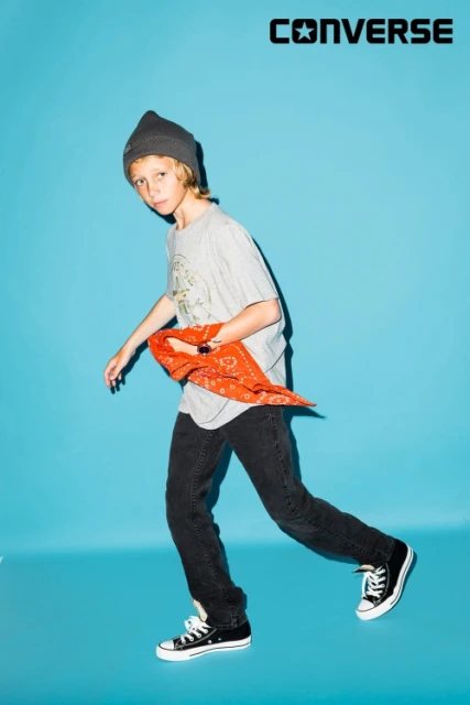 Full-length studio shot of a young boy in a t-shirt, jeans, and Converse sneakers holding a red bandana in front of a blue background.