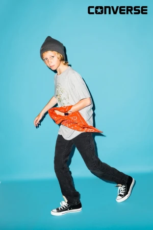 Full-length studio shot of a young boy in a t-shirt, jeans, and Converse sneakers holding a red bandana in front of a blue background.
