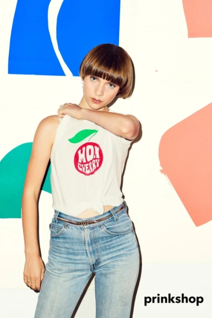 A young woman poses in a white tank top and blue jeans against a colorfully painted backdrop. Fashion and lifestyle portrait.