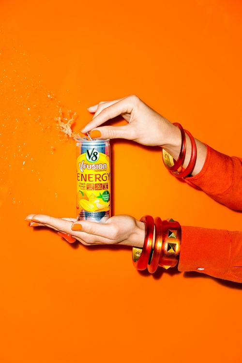Hands open a V8 V-Fusion Energy drink can, creating splash against an orange background, accessorized with orange bracelets and nails.