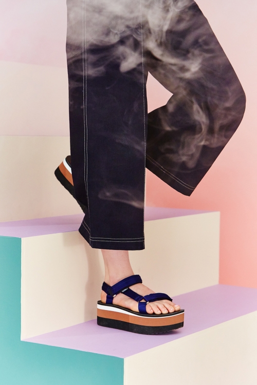 Model wears sandals and black pants on pastel stairs, overlaid with smoky effects. A fashion-forward studio shot with a modern aesthetic.