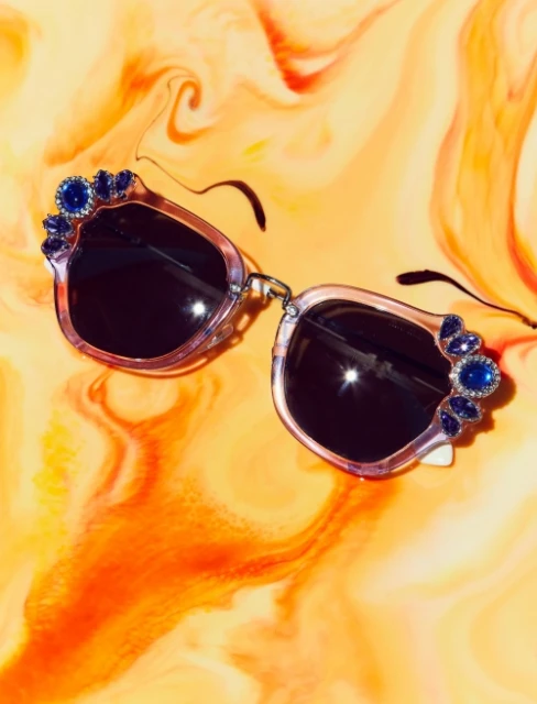 Close-up studio shot of transparent sunglasses with blue jewel embellishments on a orange, white and red swirl background. 