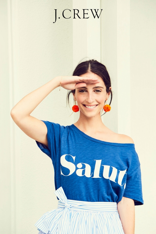 Smiling woman in a blue graphic tee and striped skirt poses with hand shielding eyes; she's wearing orange pom earrings; 'J. Crew' logo visible.