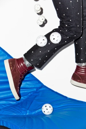 Legs wearing black and white polka-dot pants and burgundy hi-top sneakers jumping over a blue inflatable with several white perforated balls.
