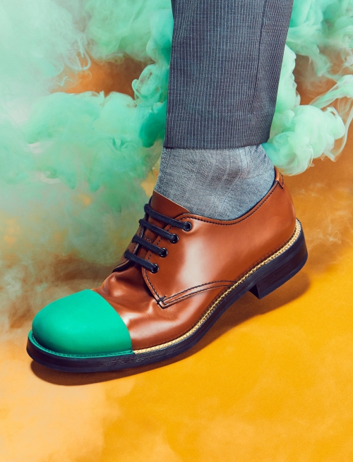 Brown leather dress shoe with a green toe cap on a gray sock and partially visible grey pants leg, standing on a yellow surface with green smoke.