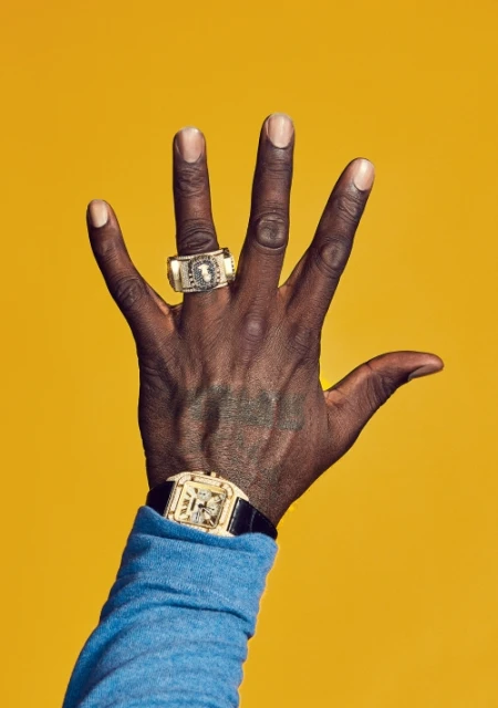 Close-up of a dark-skinned hand with a gold ring and wristwatch against a vibrant yellow backdrop. Focus on jewelry and accessories.