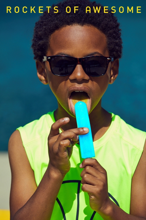 Portrait of boy with sunglasses licking blue popsicle against blue wall on sunny day wearing neon green tank top with text above.