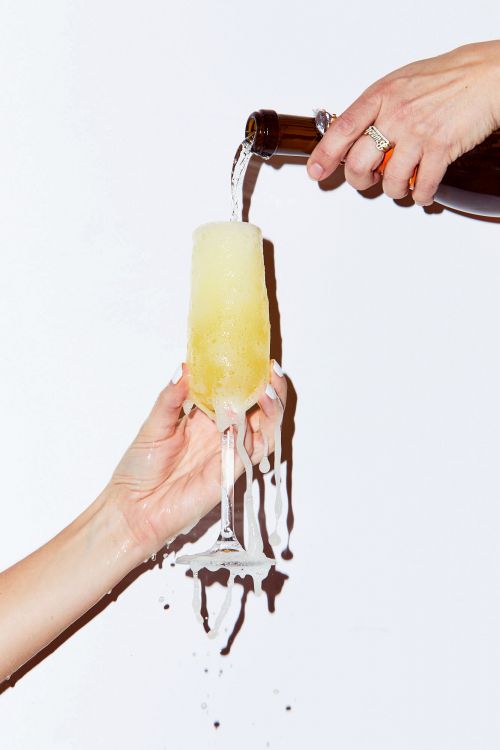 Champagne is poured into a flute, overflowing onto manicured hands, creating a visually striking celebration scene against a white background.