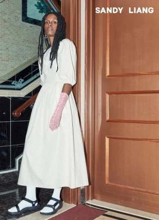 Model in white dress with pink lace gloves and sandals standing in a tiled hallway by a wooden door