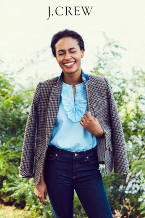 Smiling woman wearing a tweed blazer, light blue ruffled blouse, and dark denim jeans outdoors.