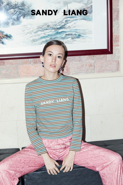 Model seated wearing a striped long sleeve top and pink trousers beneath a framed artwork.