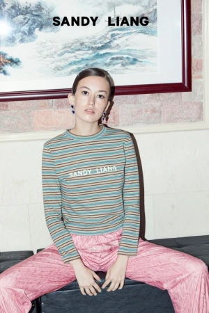 Model seated wearing a striped long sleeve top and pink trousers beneath a framed artwork.