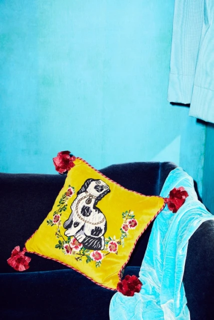 Embroidered yellow velvet cushion with a dog and floral design on a dark blue couch next to an aqua colored textile.