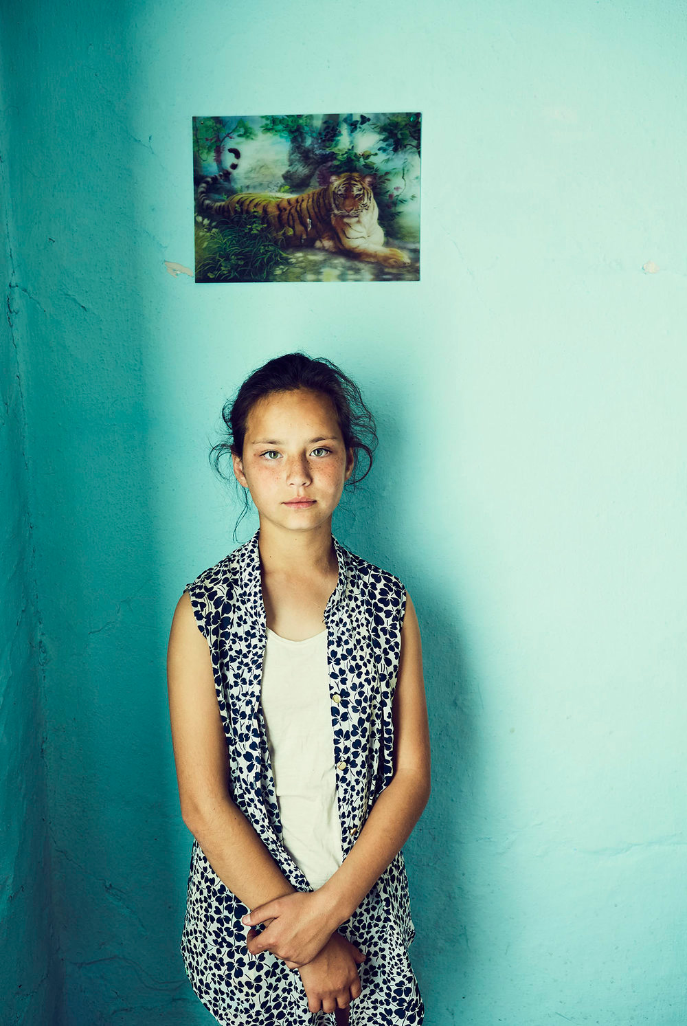 Portrait of a young girl indoors against a light blue wall, wearing a patterned top, with a tiger picture visible on the wall.