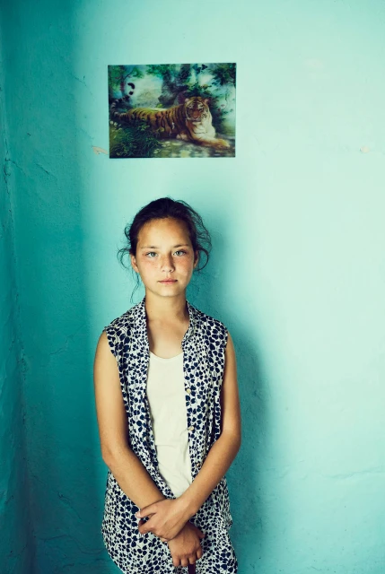 Portrait of a young girl indoors against a light blue wall, wearing a patterned top, with a tiger picture visible on the wall.