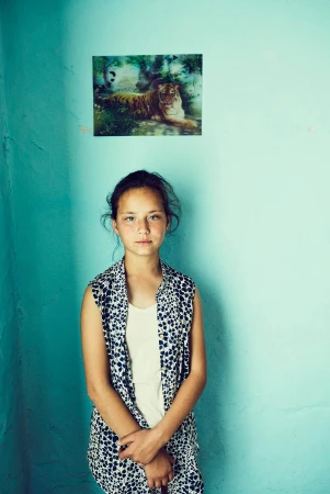Portrait of a young girl indoors against a light blue wall, wearing a patterned top, with a tiger picture visible on the wall.