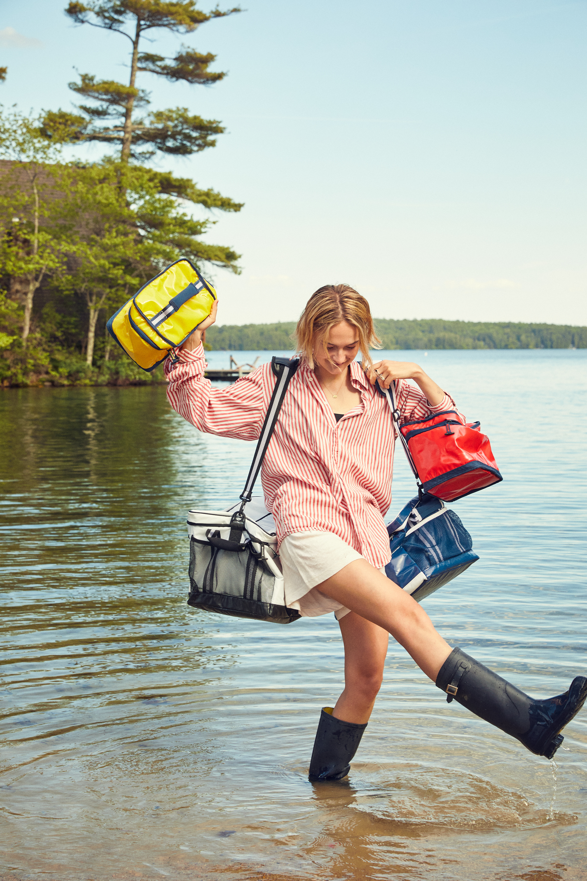 A blonde woman is wading in shallow lake water holding multiple colorful totes, while wearing a red striped shirt, white shorts, and black boots.