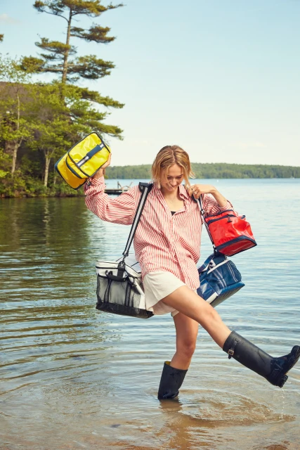 A blonde woman is wading in shallow lake water holding multiple colorful totes, while wearing a red striped shirt, white shorts, and black boots.