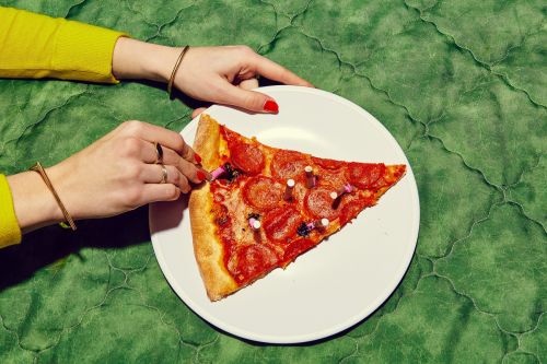 Overhead shot of a slice of pepperoni pizza on a white plate being prepared with birthday candles by a person with manicured red nails.