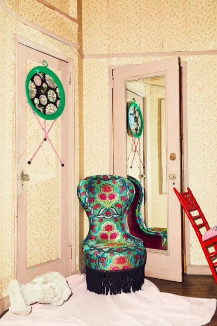 Patterned chair in room with floral wallpaper, surrounded by a pink-framed mirror and a table attached to a pink door.