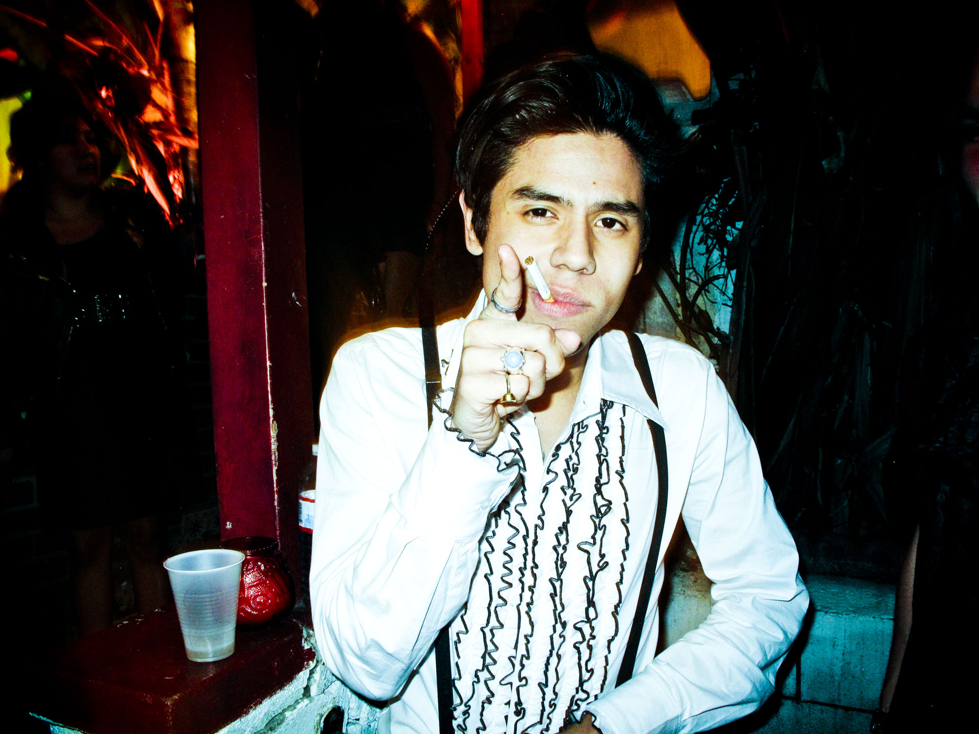 Young man in a white ruffled shirt and suspenders pointing forward with a cigarette in his mouth by photographer Meredith Jenks.