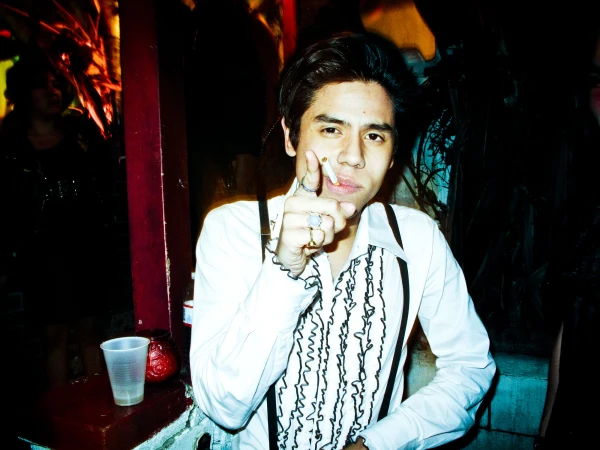 Young man in a white ruffled shirt and suspenders pointing forward with a cigarette in his mouth by photographer Meredith Jenks.