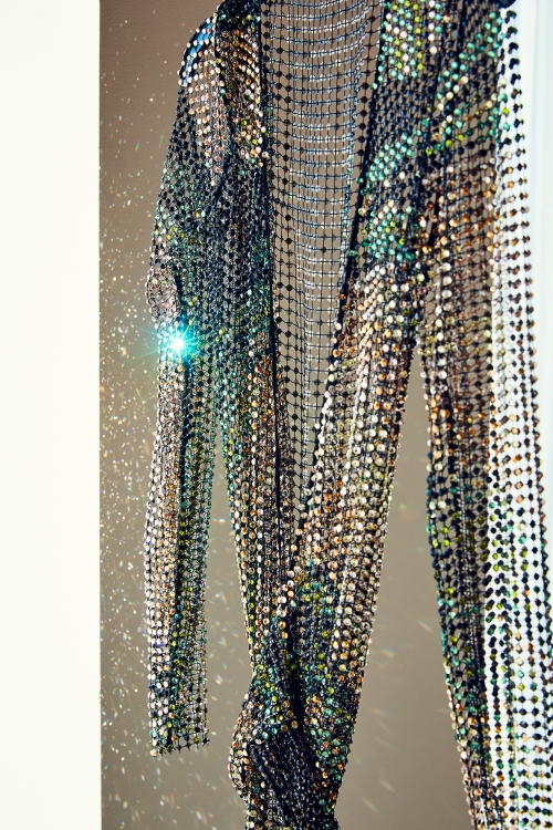 Elegant see-through dress featuring an elaborate design of shiny sequins, hanging against a neutral-toned wall with subtle shadows and light.