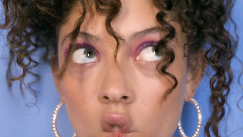 Close-up portrait of young woman with curly brown hair rolling her eyes and pursing lips against blue background.