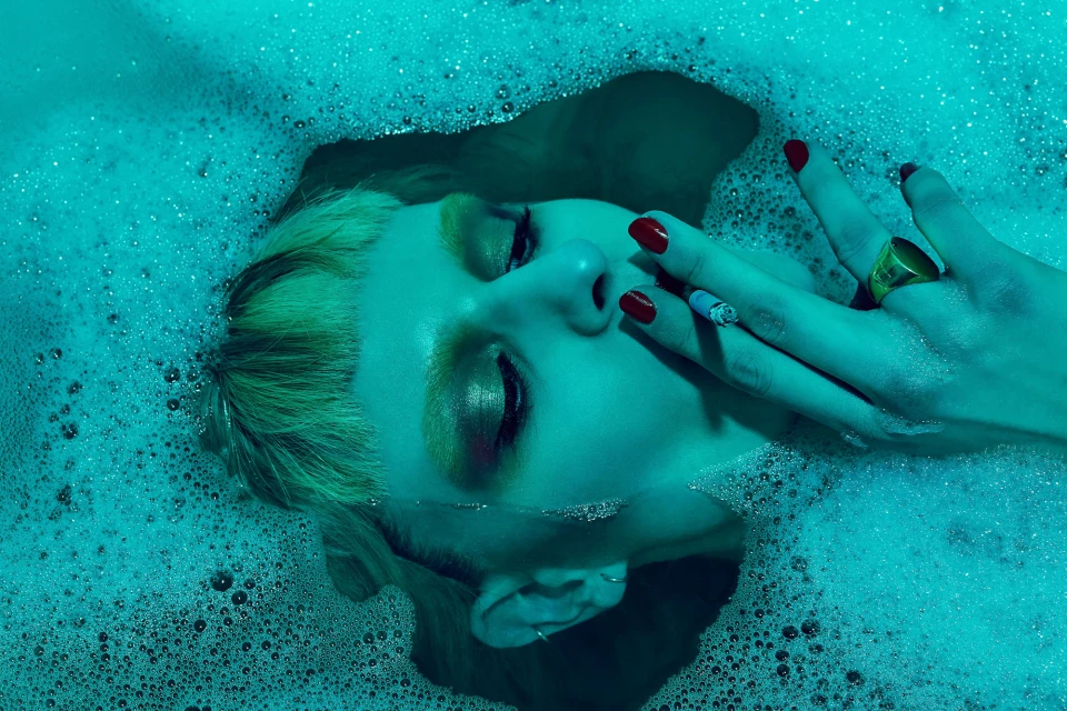 Woman lying in a tub filled with bubbly water, holding a cigarette, wearing a ring and red nail polish in blue lighting.