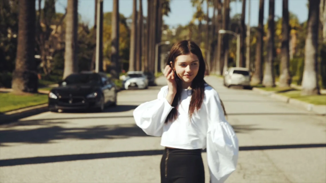 Young woman in white blouse stands on palm-lined street with luxury cars. Sunny day, lifestyle, fashion editorial, outdoor setting.