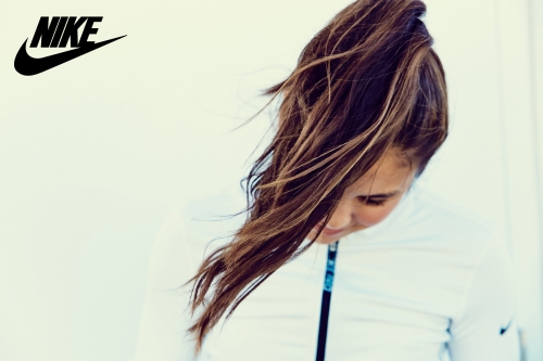 Woman with long brunette hair in a high ponytail, wearing a white jacket and facing down, against a white background with a Nike logo.