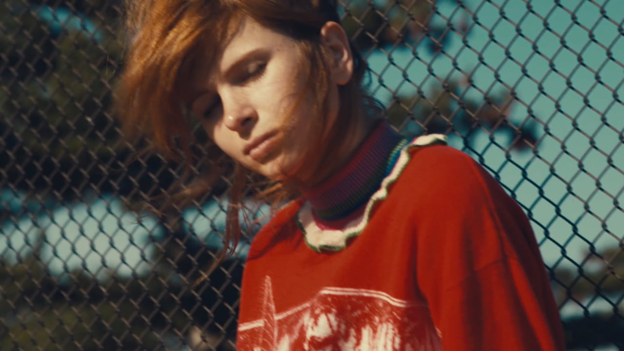 A young adult female poses outdoors with a chain-link fence behind her, wearing a red sweater with a printed design and a colorful ruffled collar.