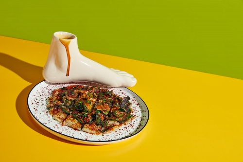 Still life food photo: savory pancake on a plate, next to a ceramic foot-shaped container dripping with brown sauce, against yellow-green background.